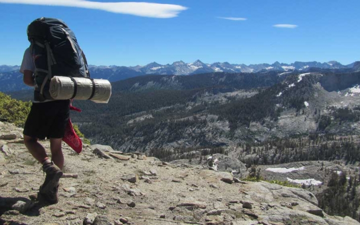 a person wearing a backpack hikes along rocky terrain high above a mountainous landscape